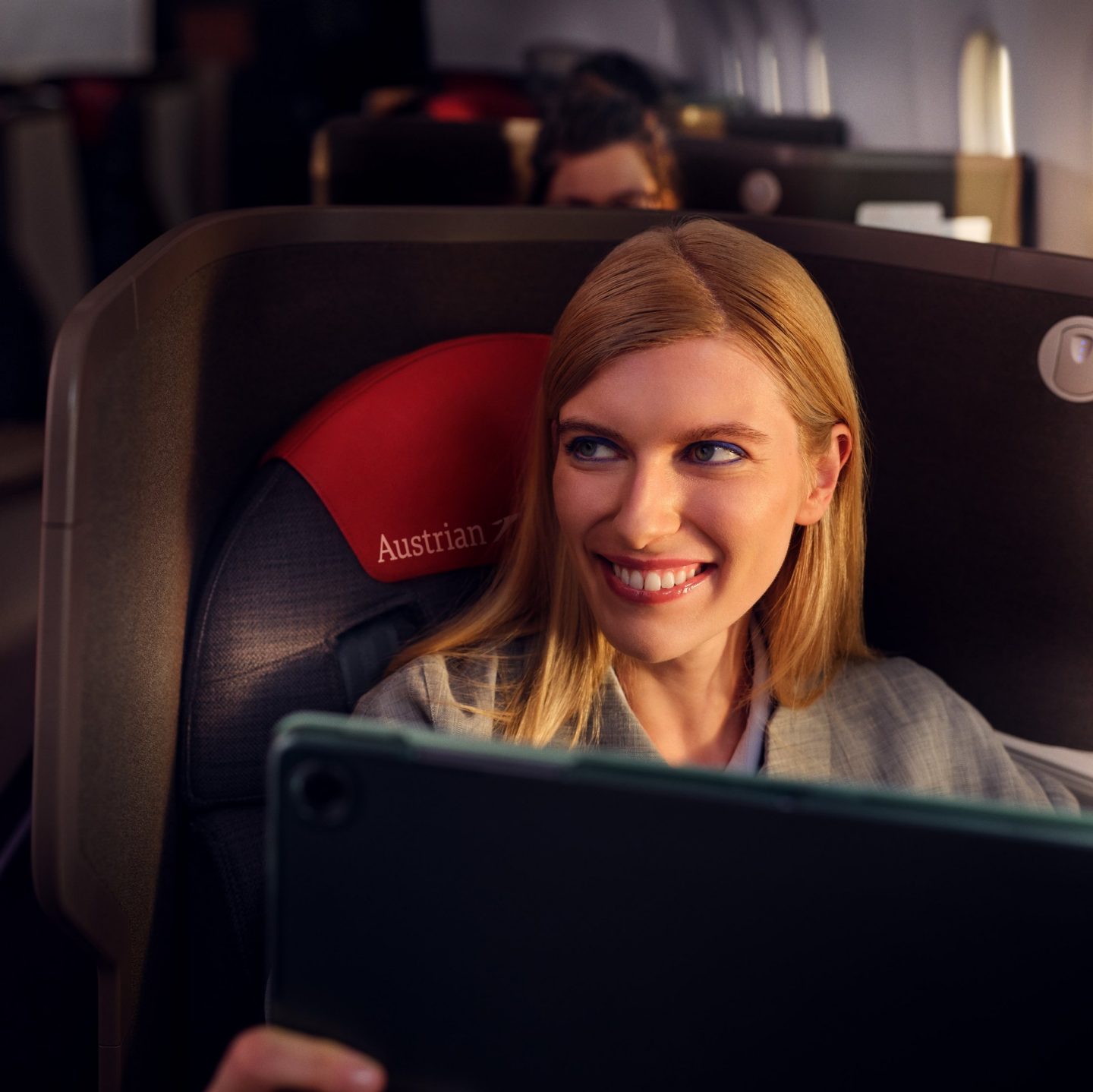 A smiling blonde woman sitting in an Austrian Airlines seat, looking at a tablet.