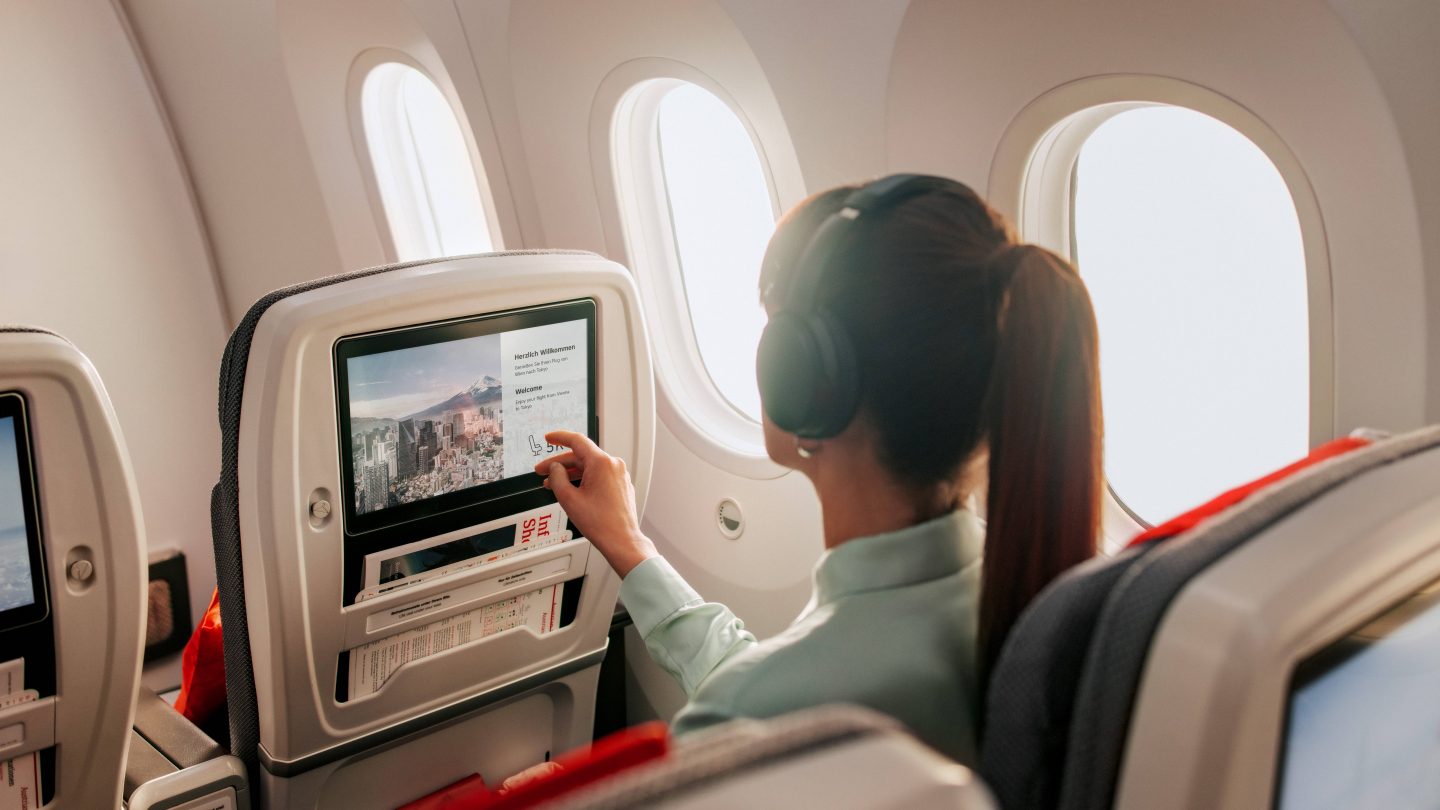 A woman wearing headphones taps an airplane seatback entertainment screen beside bright windows, appearing relaxed during a calm flight.