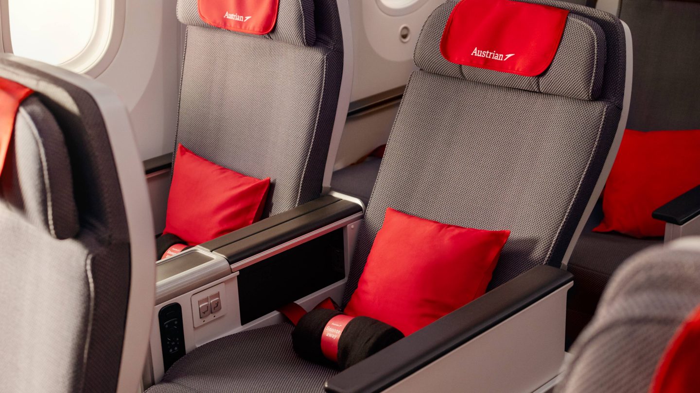Rows of grey airplane seats with red headrest covers, pillows, and Austrian Airlines branding.