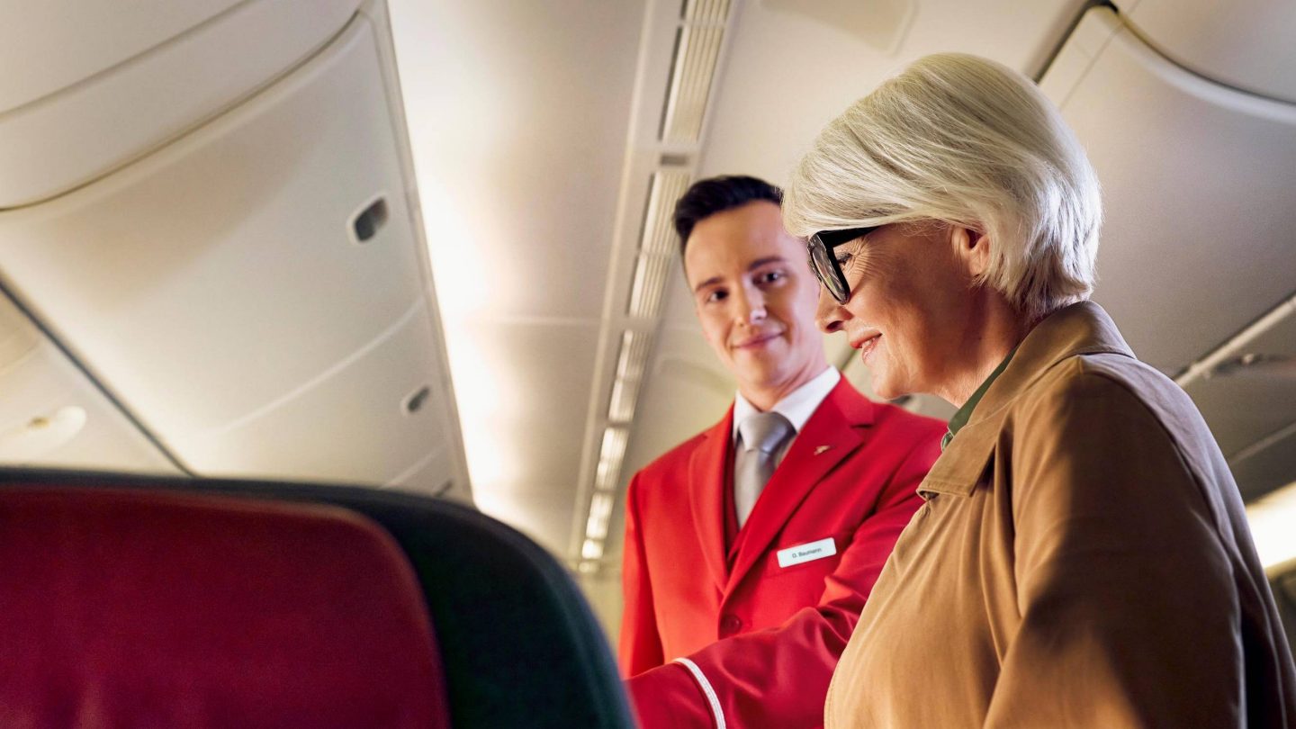 A flight attendant in a red uniform helps a female passenger to her seat on an airplane.