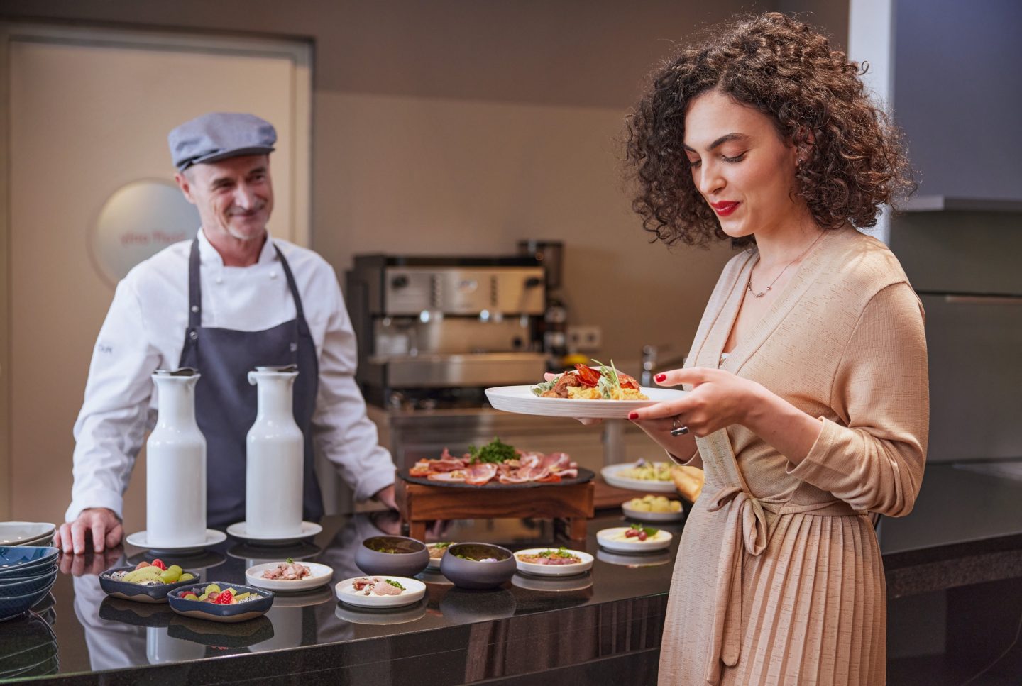 A woman selecting food from a buffet while a chef watches in the background.