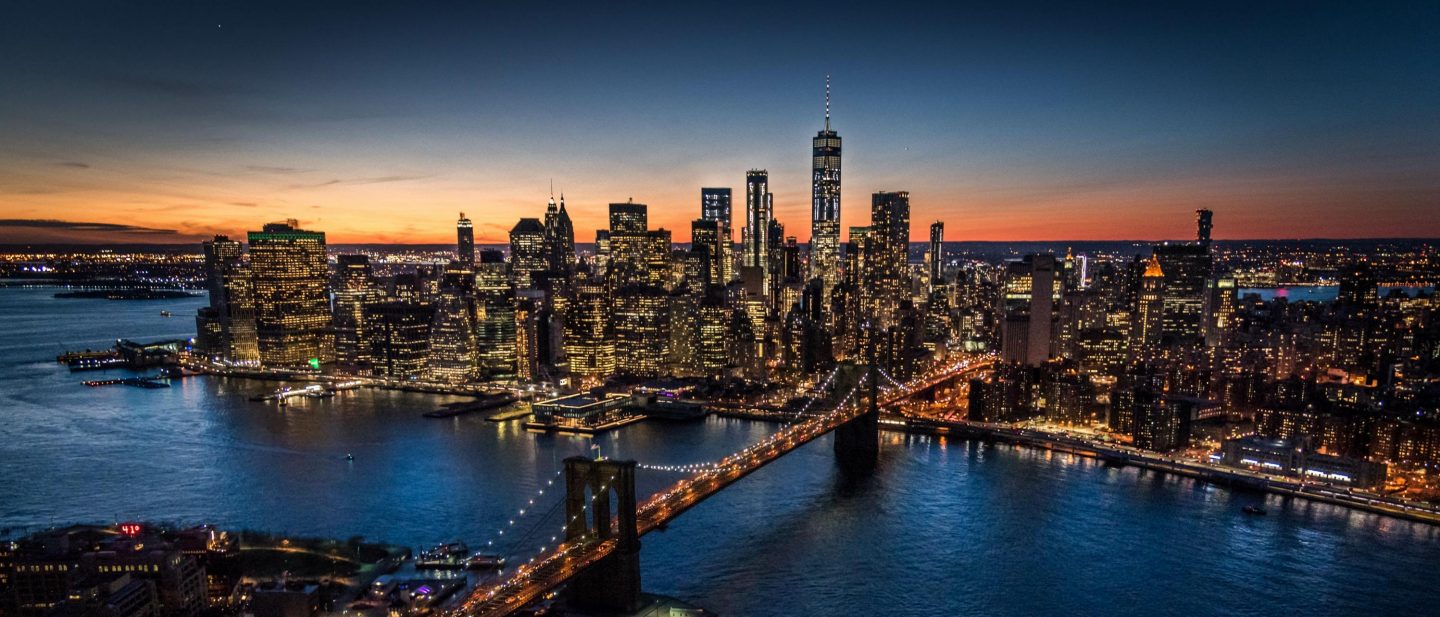 Night view of New York City with the illuminated Brooklyn Bridge leading to the Manhattan skyline under a vibrant sunset sky.