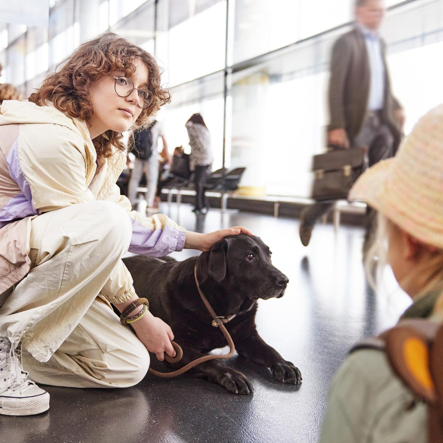 Person with a dog sitting in an airport terminal while travelers pass by. In the foreground, a child with a backpack is looking at the dog.