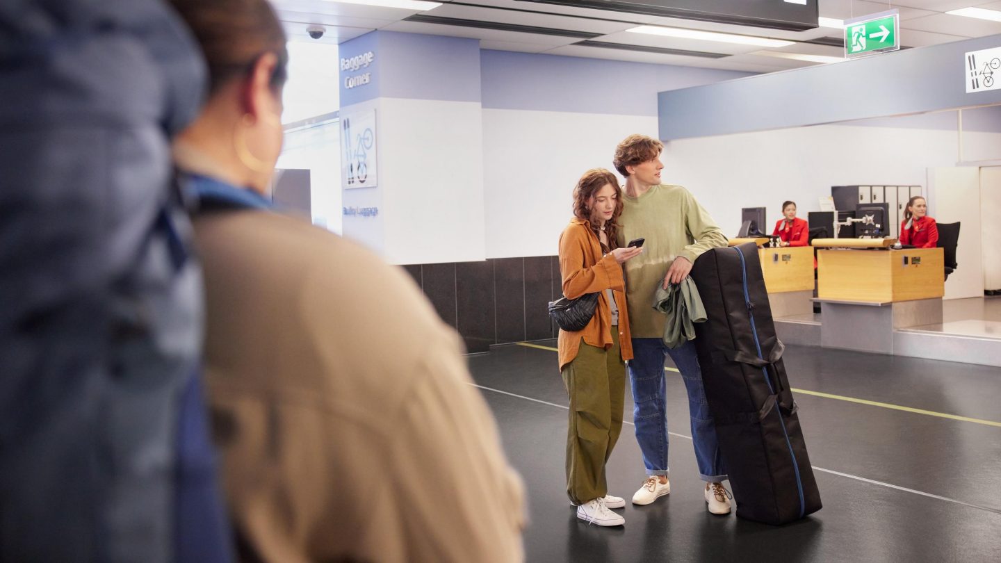 Man walking in an airport with a large musical instrument case, surrounded by travelers.