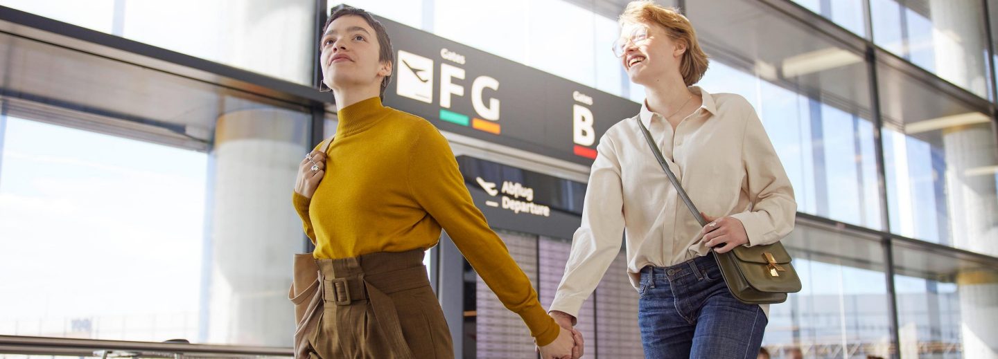 Two women holding hands walking in an airport terminal near departure gates FG and B signs.