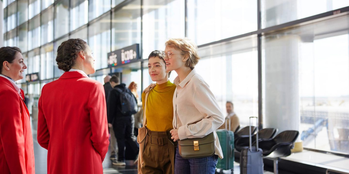 Smiling airline staff greet two happy passengers in a bright airport terminal.