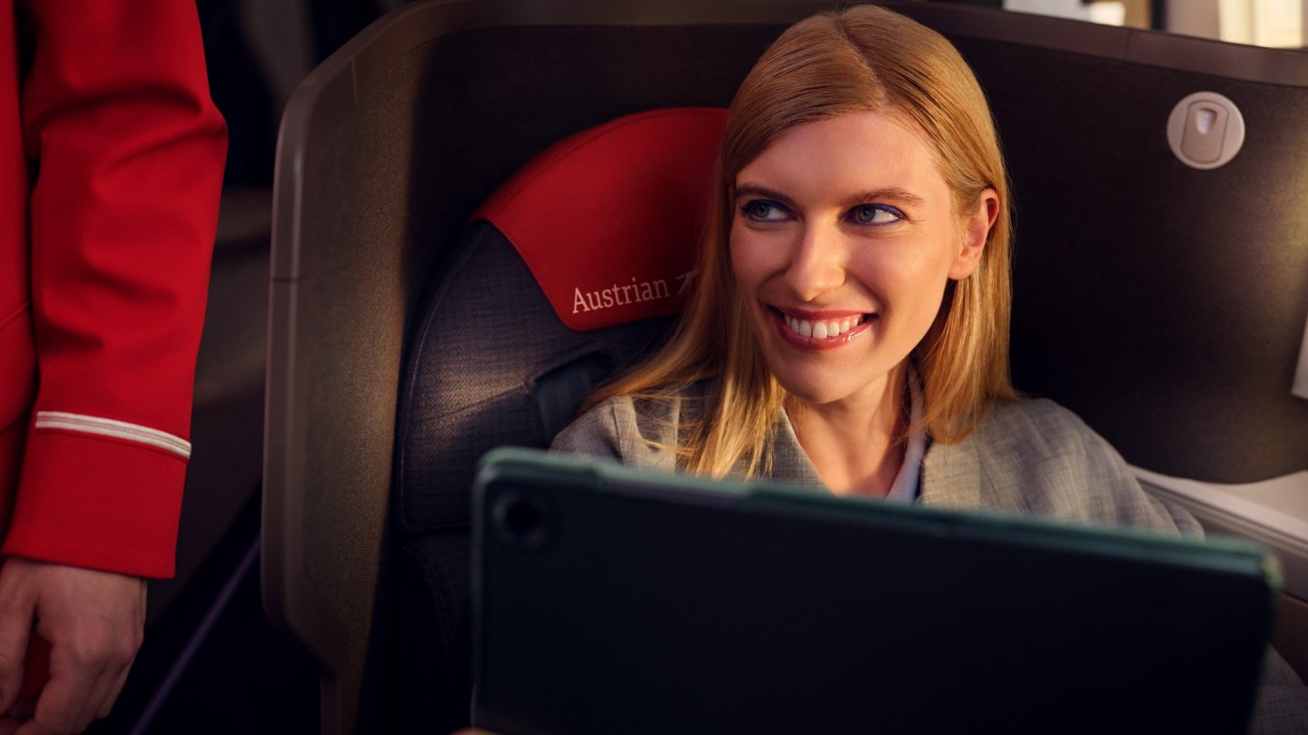 A smiling blonde woman sitting in an Austrian Airlines seat, looking at a tablet.