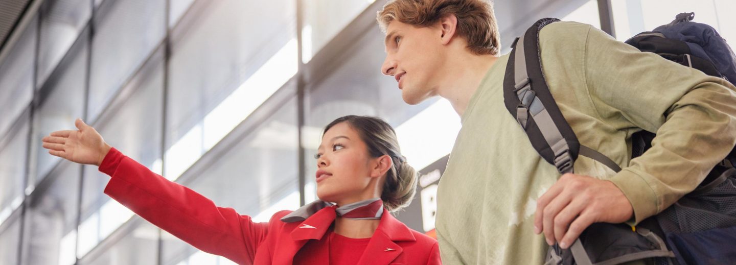 A flight attendant in red uniform points the way for a traveler with a backpack in an airport.