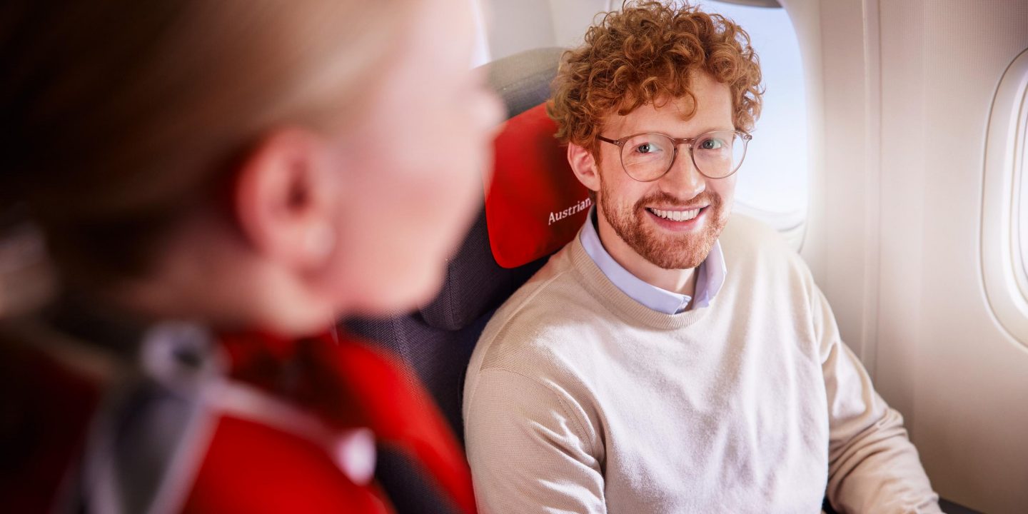 A smiling man with red curls and glasses sits comfortably in an airplane seat.