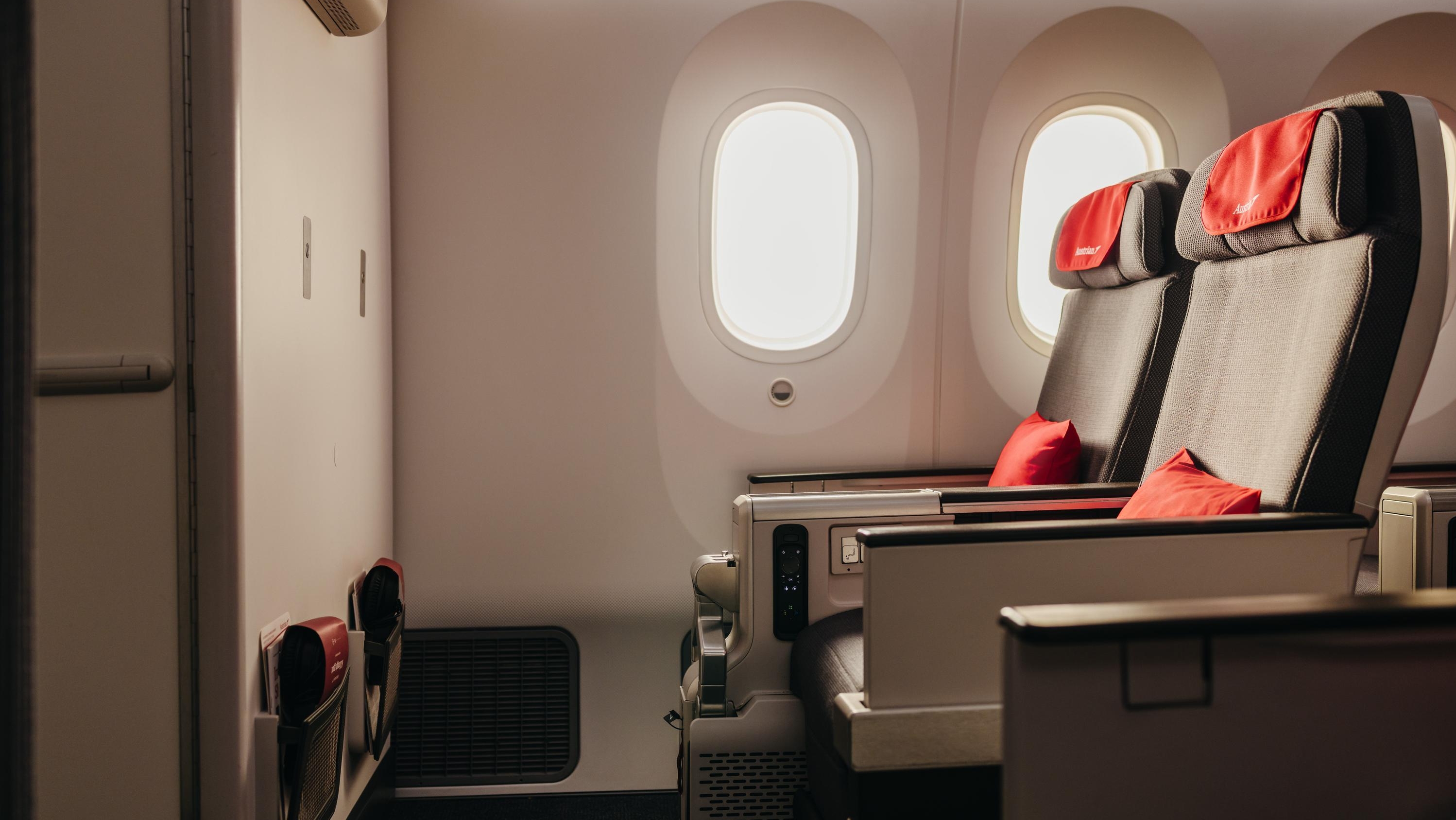 Spacious airplane cabin with two wide seats, red headrests and pillows, softly lit by two oval windows.