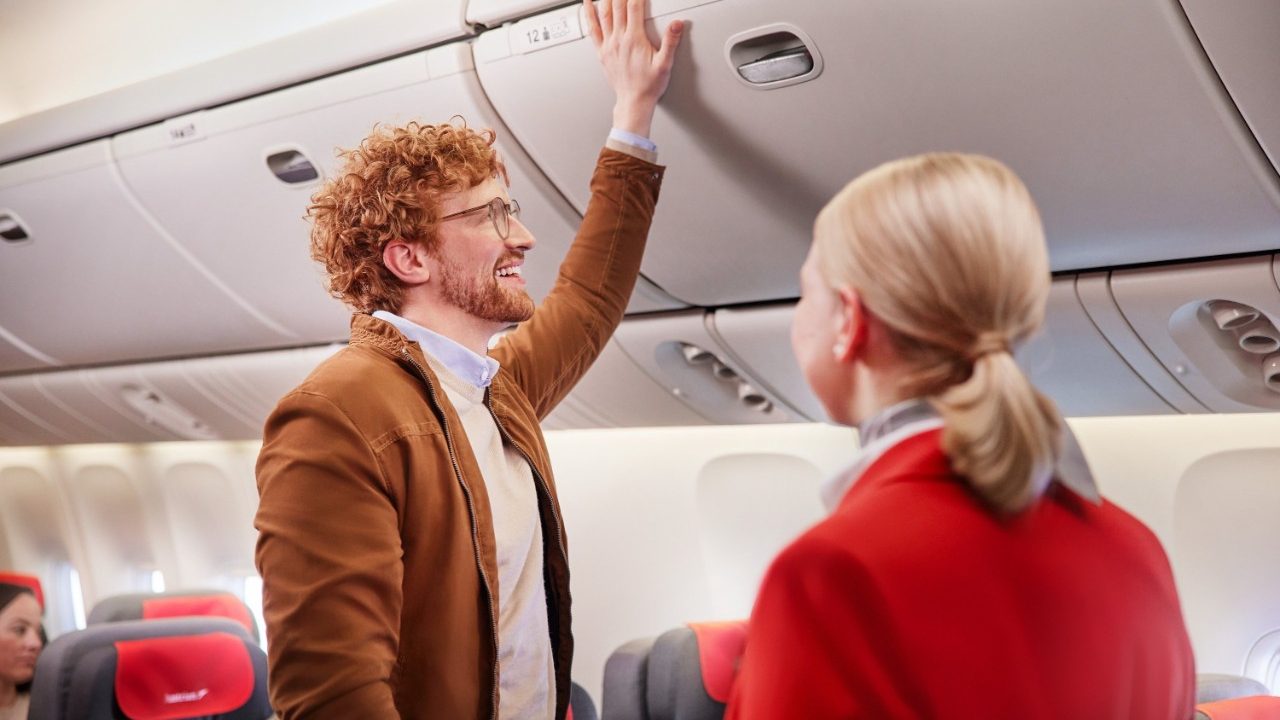 Smiling passenger reaching for an overhead bin while a flight attendant looks on.