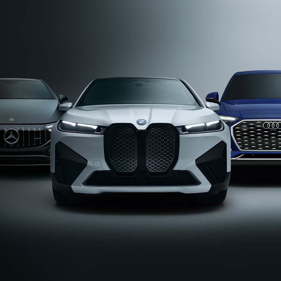 Front view of three cars from different brands against a dark background: a black Mercedes on the left, a white BMW in the center, and a blue Audi on the right.
