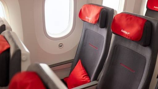 Rows of airplane seats with dark grey upholstery and red headrests featuring the Austrian Airlines logo.