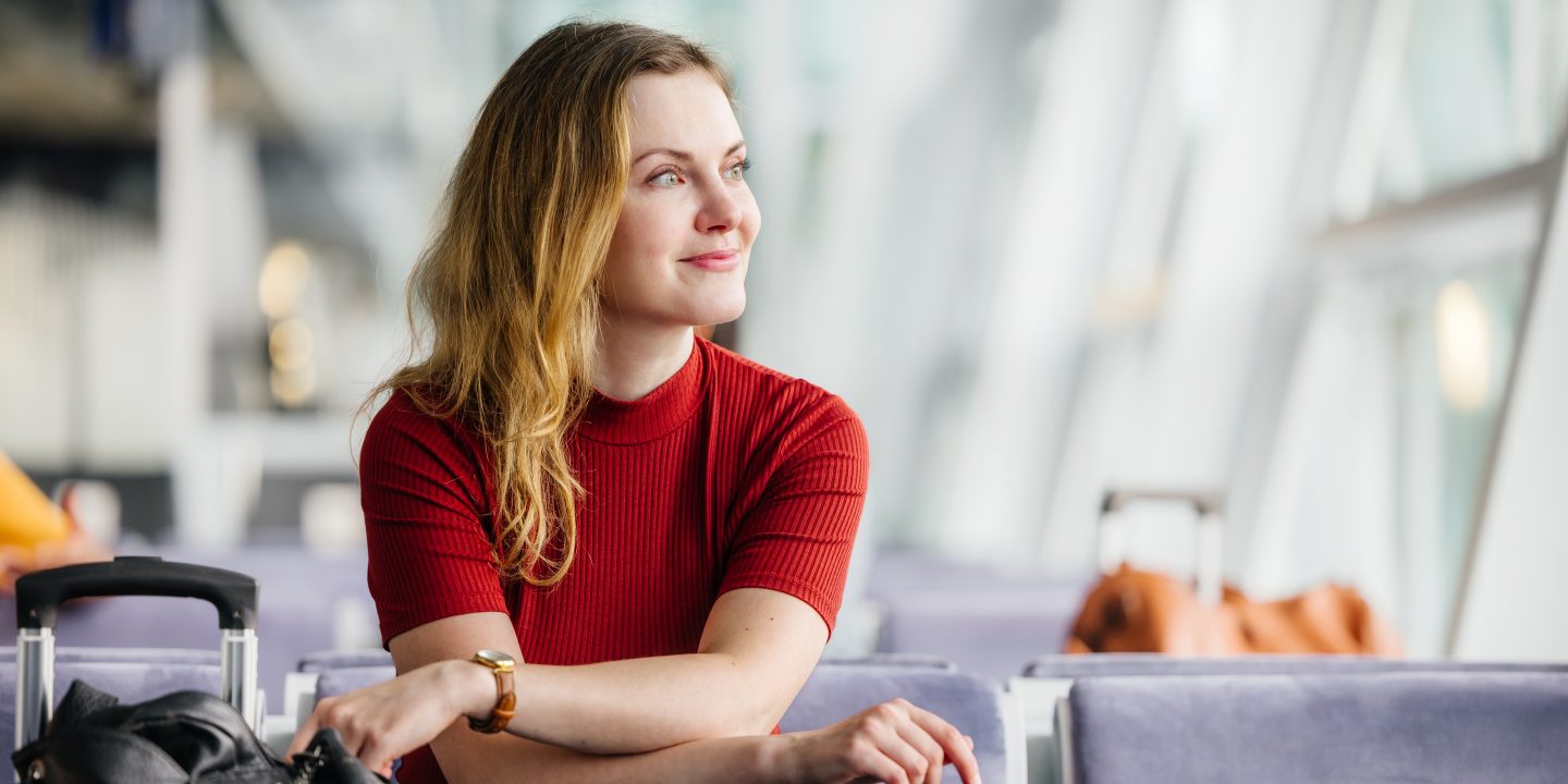An elegant woman with strawberry-blonde hair smiles while sitting next to her luggage, looking out the window and waiting for her flight in an airport lounge.