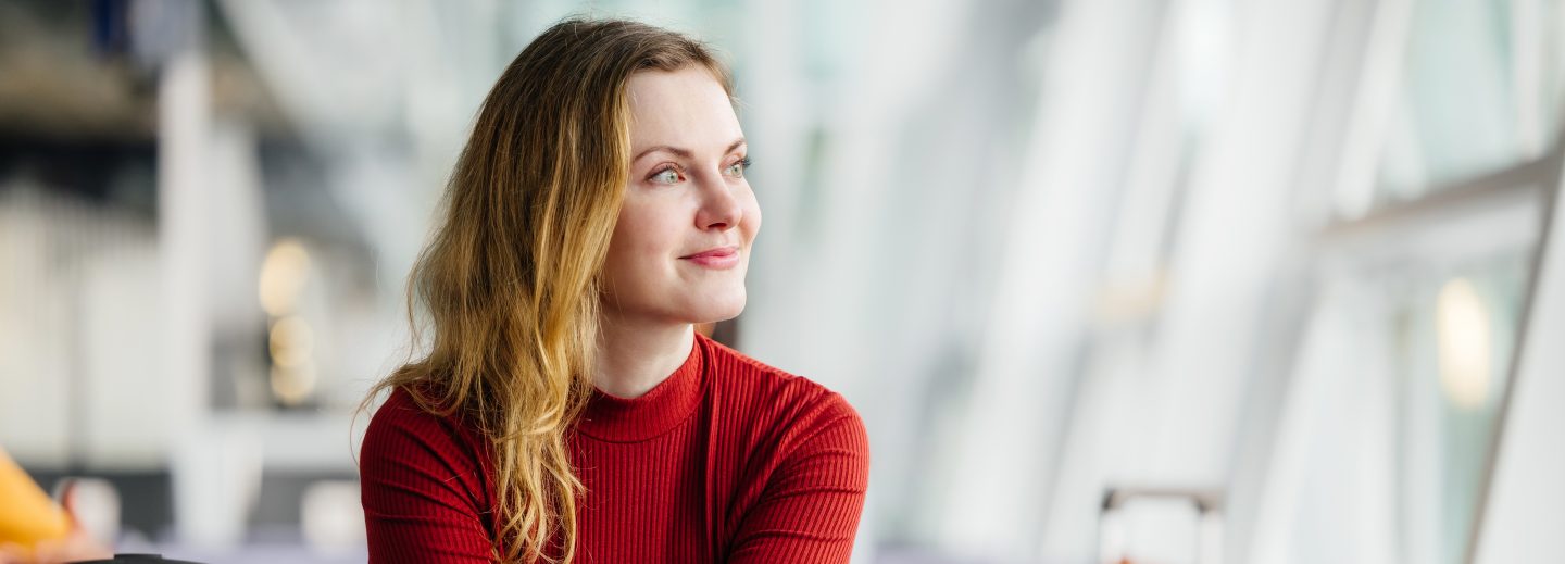 An elegant woman with strawberry-blonde hair smiles while sitting next to her luggage, looking out the window and waiting for her flight in an airport lounge.